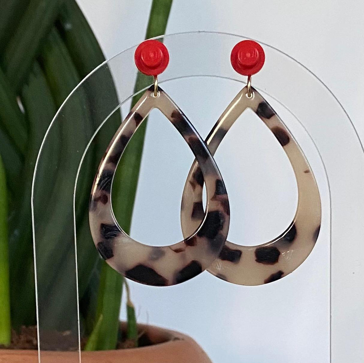 Boucles d’oreilles raffinée et tendance en forme de goutte en acétate au motif écaille de tortue avec clou d’oreille en brique Lego rouge, présentées sur un support transparent devant un cactus.