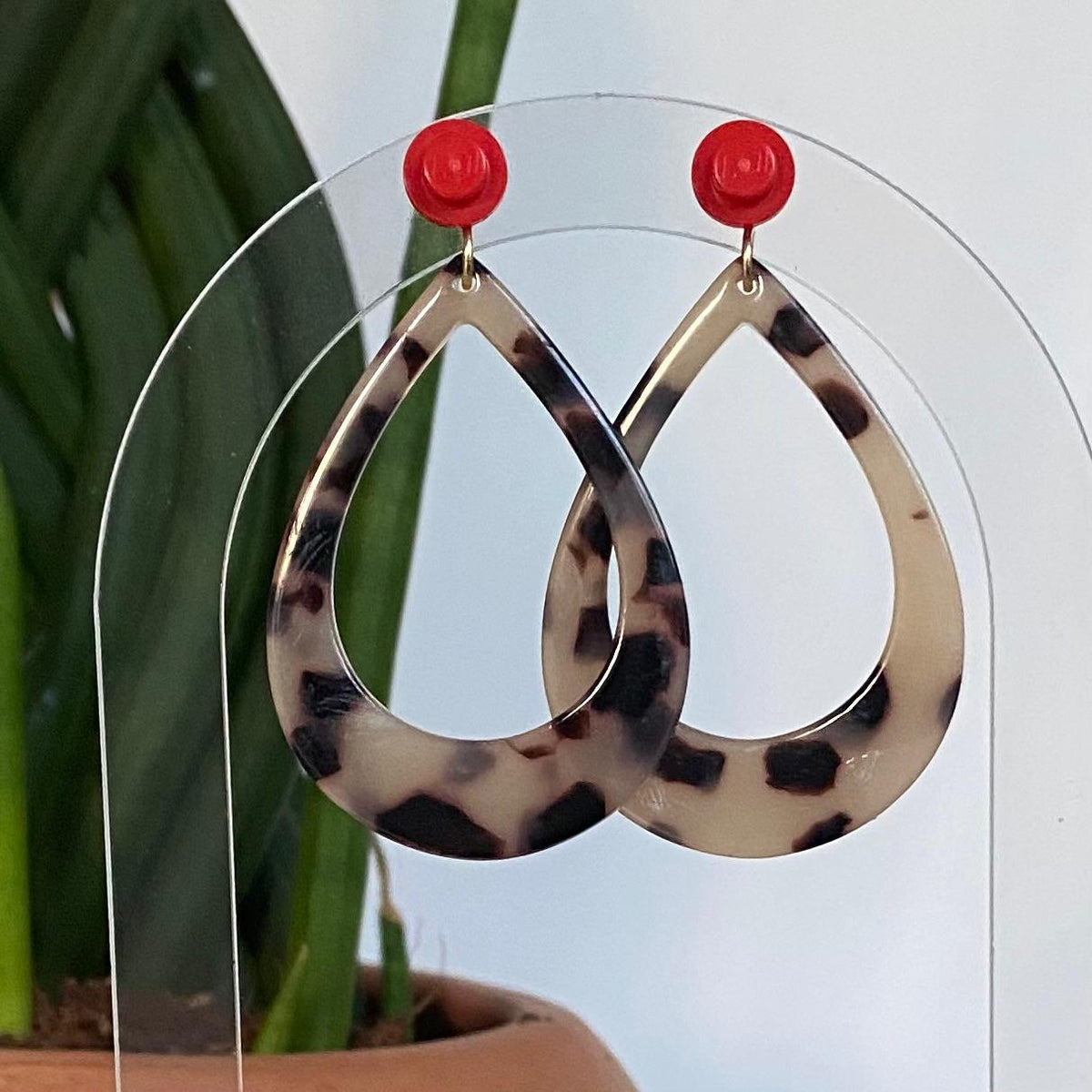 Boucles d’oreilles raffinée et tendance en forme de goutte en acétate au motif écaille de tortue avec clou d’oreille en brique Lego rouge, présentées sur un support transparent devant un cactus.