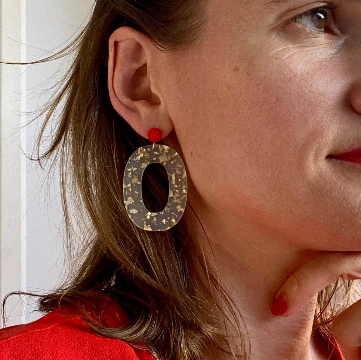 Boucles d'oreille paillettes or et Lego portées par une femme, avec une pièce ovale dorée et une brique Lego rouge.