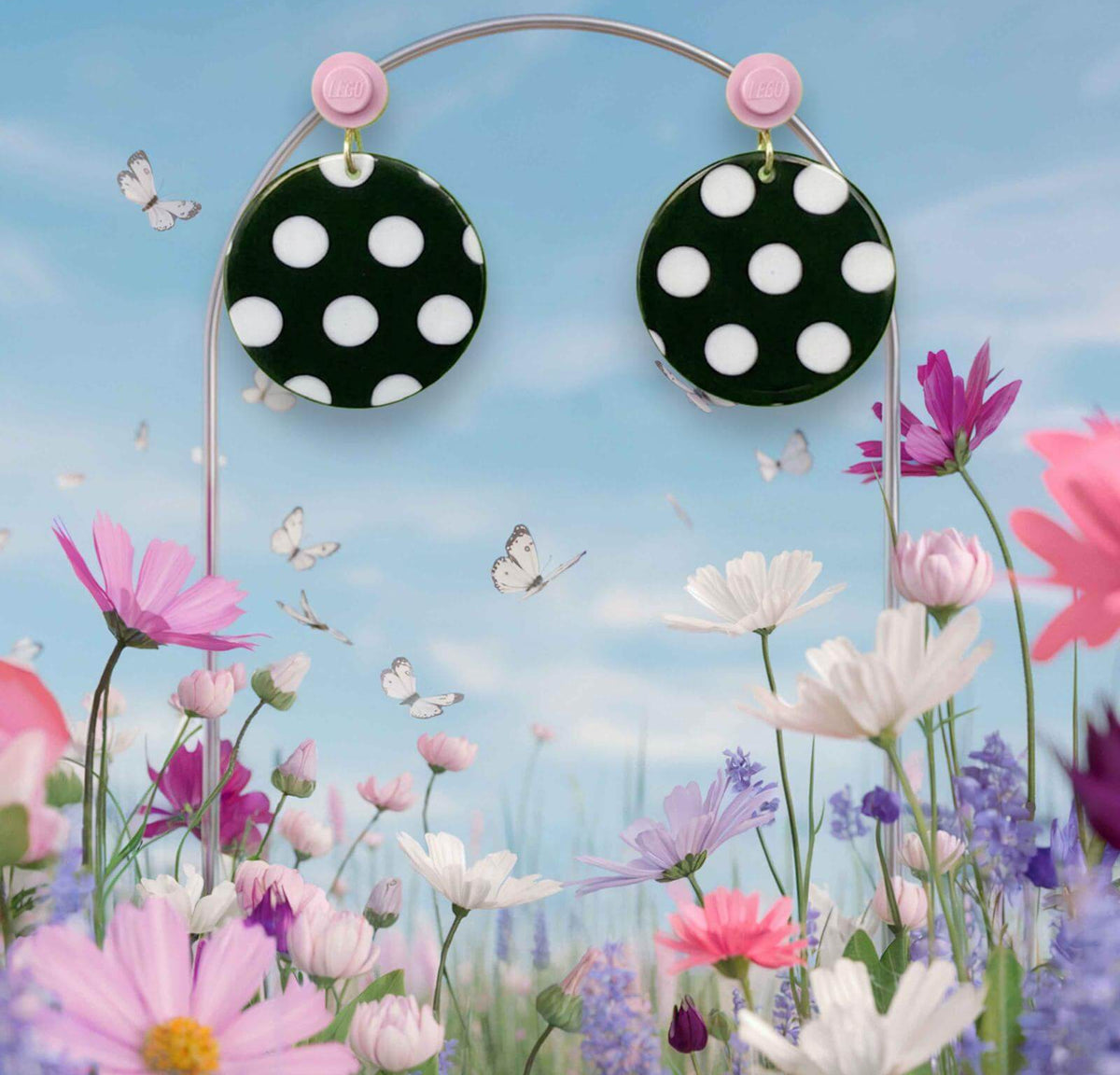 Boucles d'oreilles à pois noirs et blancs accrochées à l'oreille par une brique de Lego ronde rose pastel au milieu d'un décor féérique floral