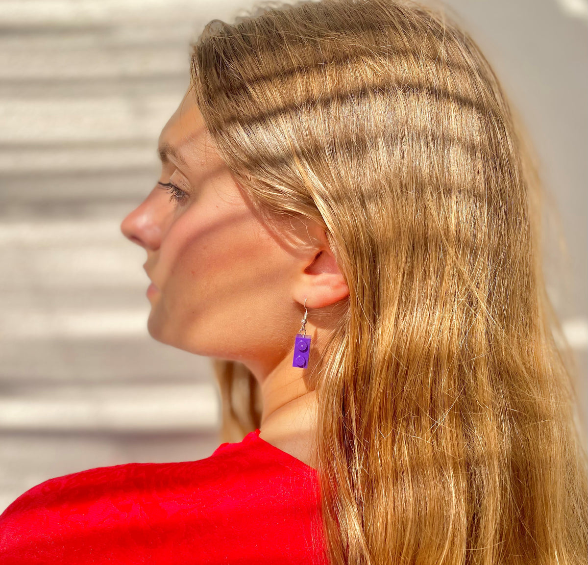 Femme portant des boucles d'oreilles pendantes en brique Lego violette, cheveux longs et blonds, vêtue d'un haut rouge.