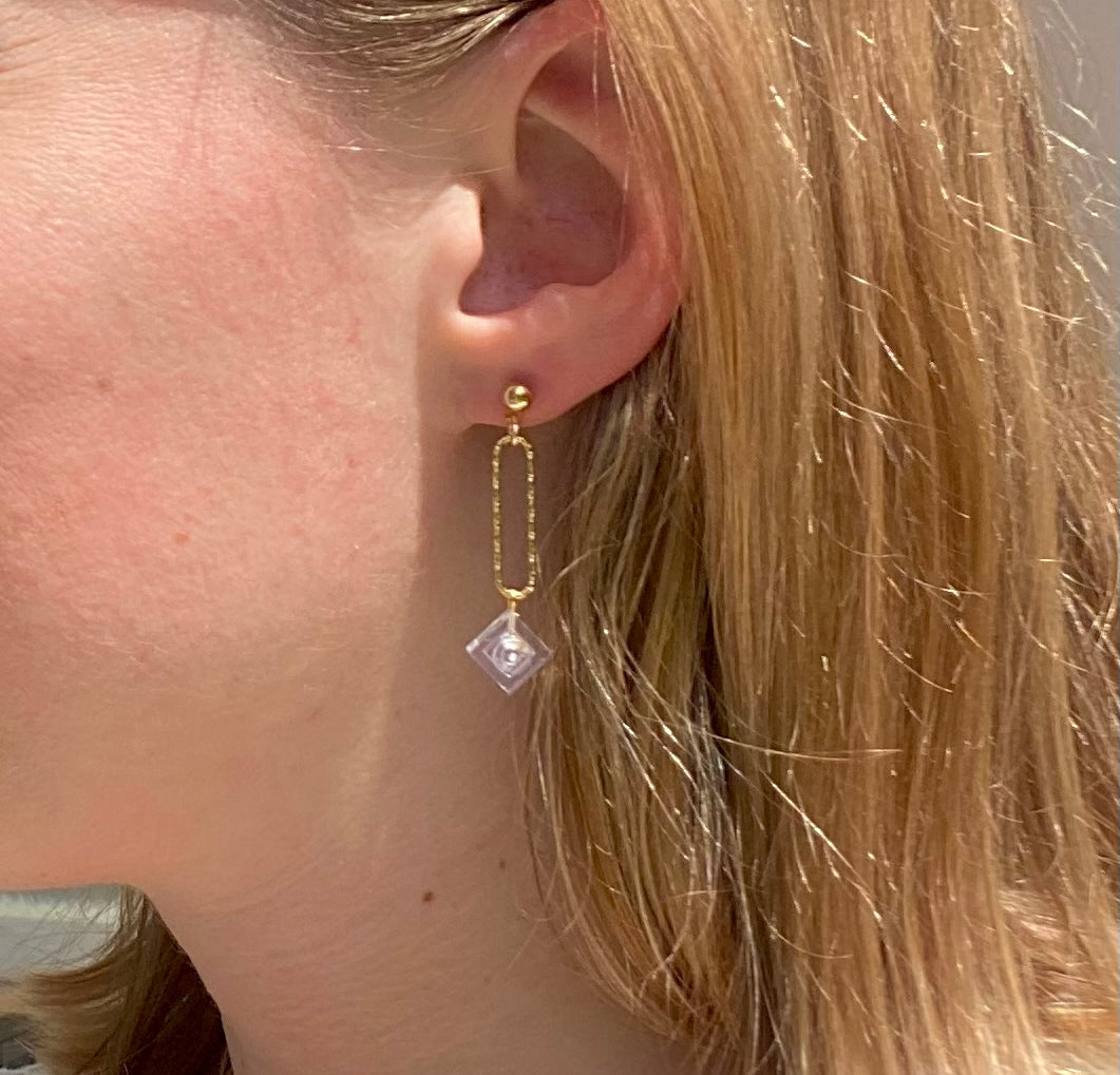 Boucle d'oreilles pendante accrochée à l'oreille par un clou d'oreille en forme de petite boule auquel est suspendu un anneau doré ovale martelé et en-dessous une brique de Lego 1x1 transparente comme du cristal. La boucle d'oreille est portée par une femme blanche blonde.