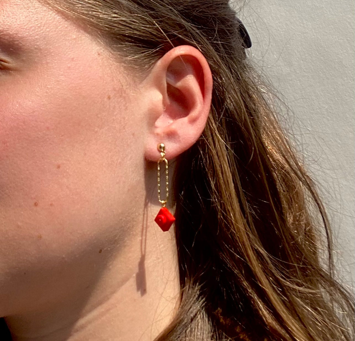 Boucle d'oreilles pendante accrochée à l'oreille par un clou d'oreille en forme de petite boule auquel est suspendu un anneau doré ovale martelé et en-dessous une brique de Lego 1x1 plate rouge vif. La boucle d'oreille est portée par une femme blanche châtain..