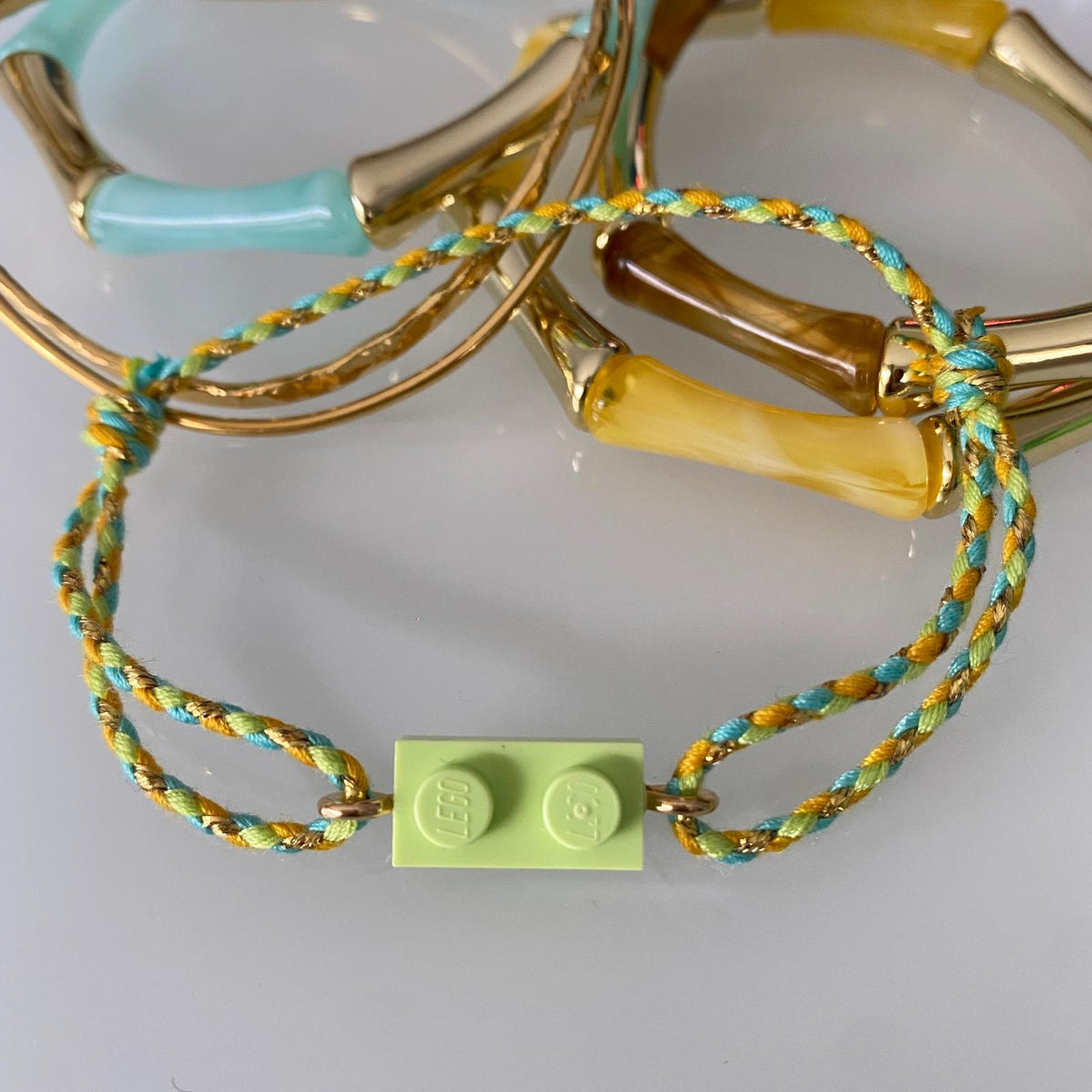 Bracelet posé à plat avec une brique de Lego et fermé par un cordon ajustable fin tressé vert pale, orangé, turquoise et doré. Il est présenté avec d'autres bracelets fantaisies tendance style bambou aux couleurs assorties.