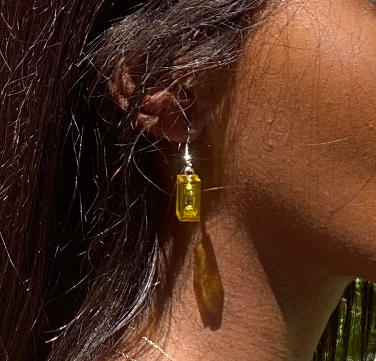 Boucles d'oreilles pendantes en brique Lego® jaune, ajoutant une touche colorée au look, portées avec des cheveux longs.