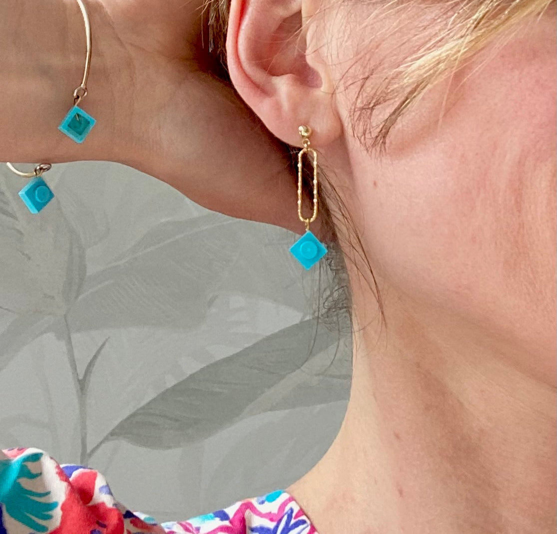 Boucle d'oreilles pendante accrochée à l'oreille par un clou d'oreille en forme de petite boule auquel est suspendu un anneau doré ovale martelé et en-dessous une brique de Lego 1x1 bleu turquoise. La boucle d'oreille est portée par une femme blanche blonde. Elle porte le bracelet Brique d'Oreille "Rockeuses" assorti.