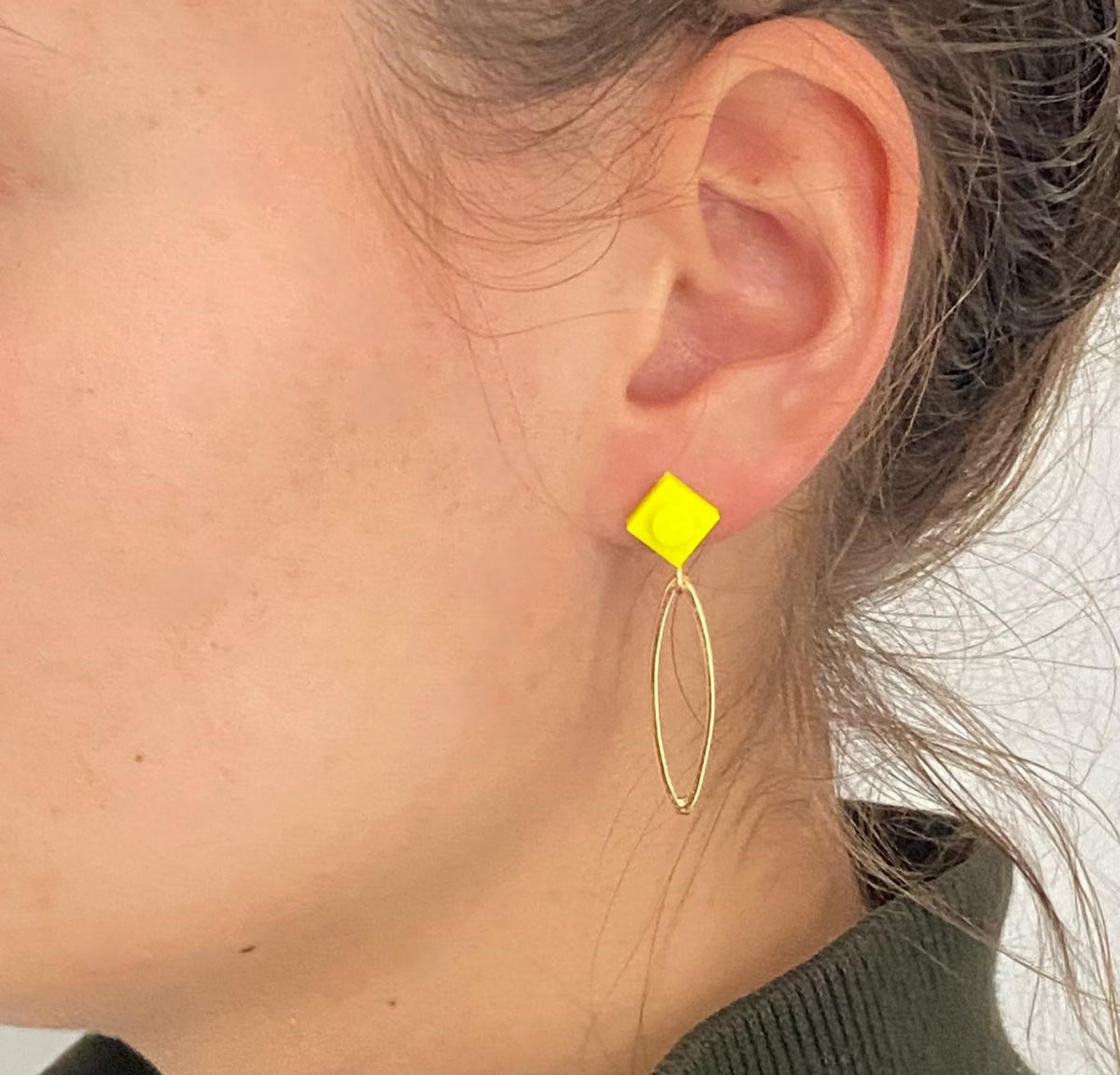 Boucle d'oreilles pendante avec un oval en laiton doré suspendue à une brique de Lego 1x1 plate jaune fluo. Elle se coordonne parfaitement avec un pull vert kaki.