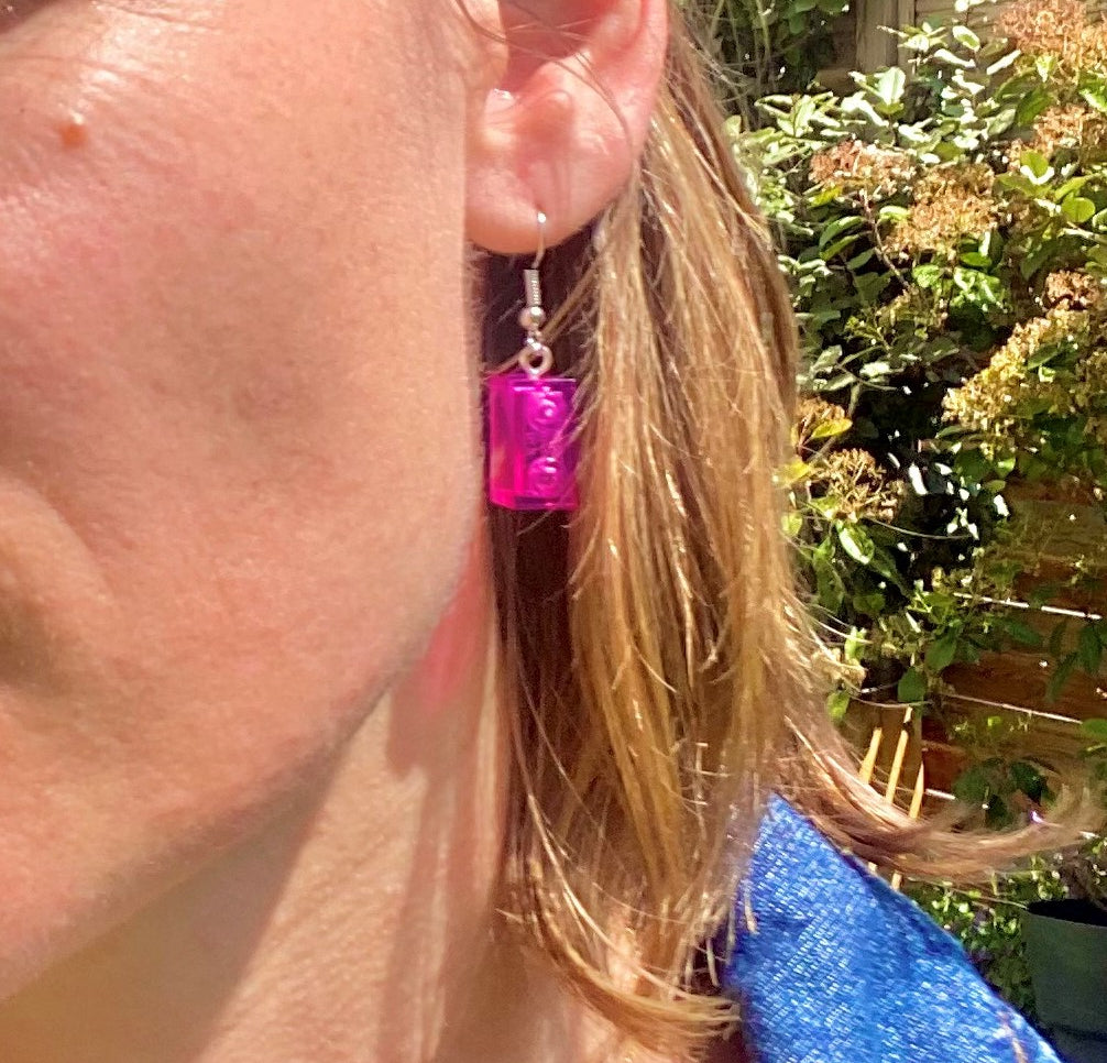 Boucles d'oreilles pendantes argentées avec brique Lego® rose accrochée à l'oreille en arrière-plan fleuri.
