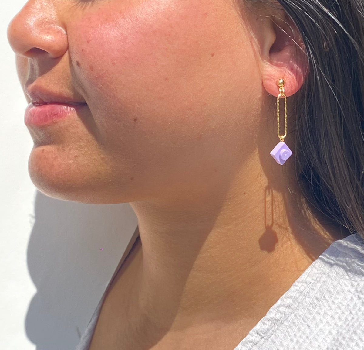 Boucle d'oreilles pendante accrochée à l'oreille par un clou d'oreille en forme de petite boule auquel est suspendu un anneau doré ovale martelé et en-dessous une brique de Lego 1x1 mauve pastel. La boucle d'oreille est portée par une femme malgache brune