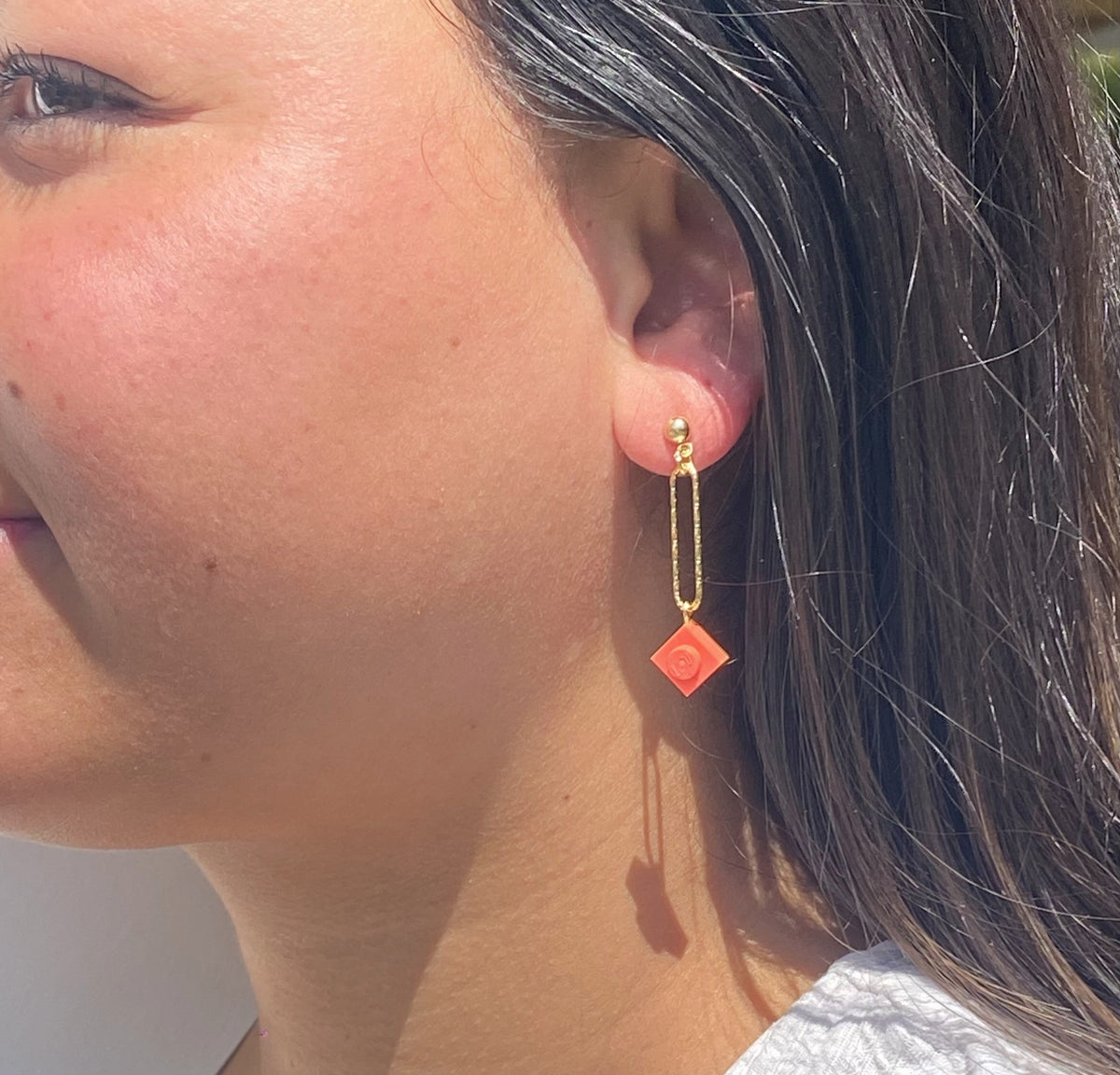 Boucle d'oreilles pendante accrochée à l'oreille par un clou d'oreille en forme de petite boule auquel est suspendu un anneau doré ovale martelé et en-dessous une brique de Lego 1x1 rose corail. La boucle d'oreille est portée par une femme malgache brune