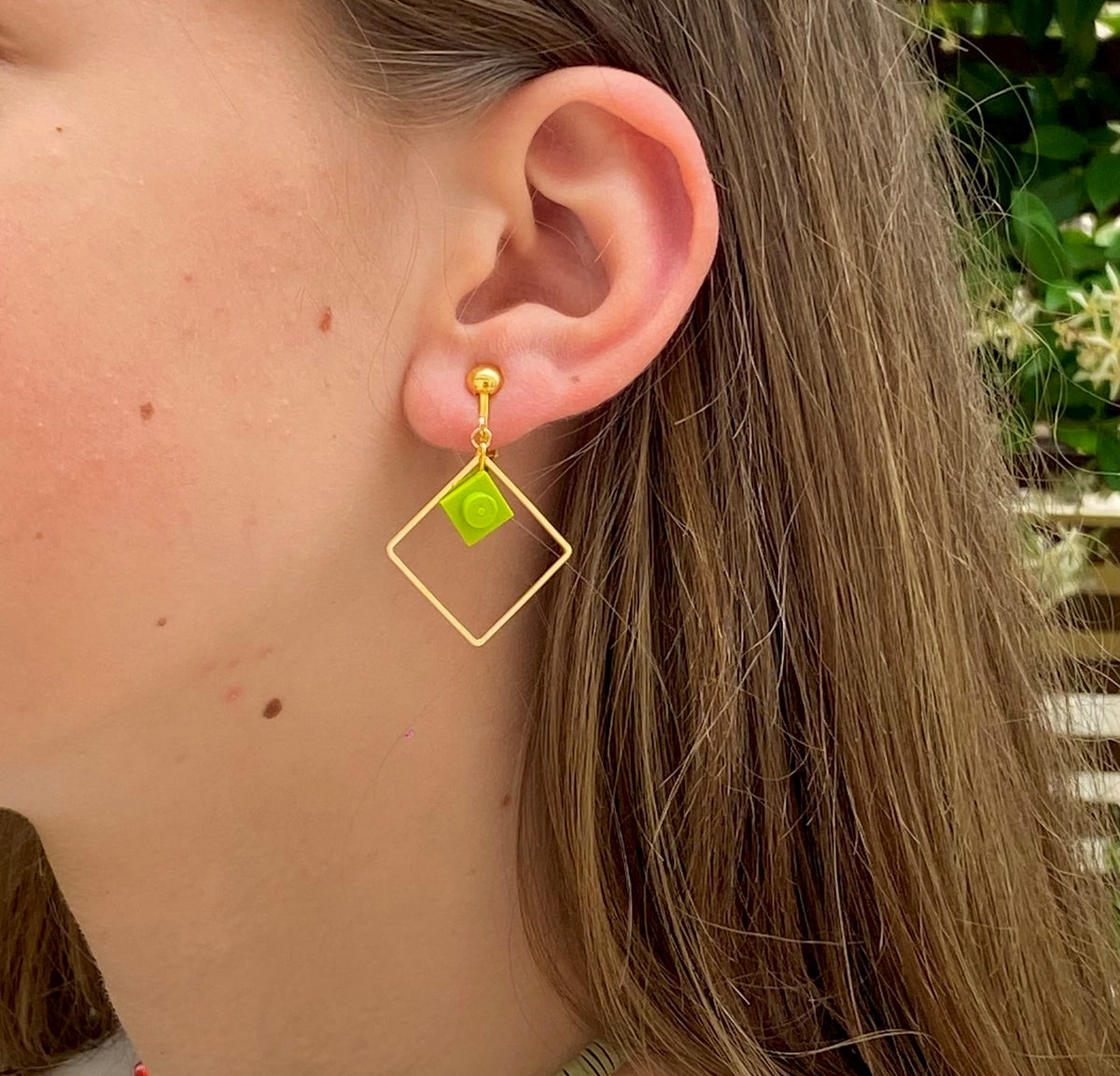 Ravissante boucle d'oreilles clip pour les oreille non percée composée d'un rectangle fin et évidé en laiton doré avec une mini brique de Lego 1x1 assemblé à l'intérieur, le tout fixé à l'oreille par un clip doré sobre. Elle est portée sur une jaune fille blonde aux cheveux longs de profil