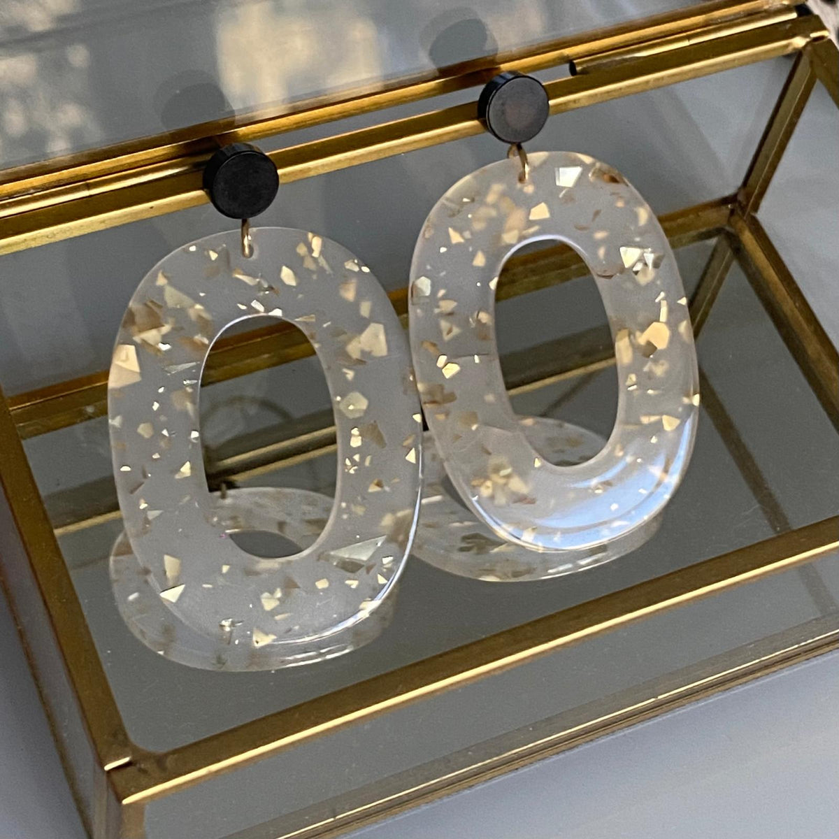 Boucles d'oreille paillettes or et Lego avec pièce ovale en acétate et éclats dorés, présentation sur une boîte à bijoux en verre.