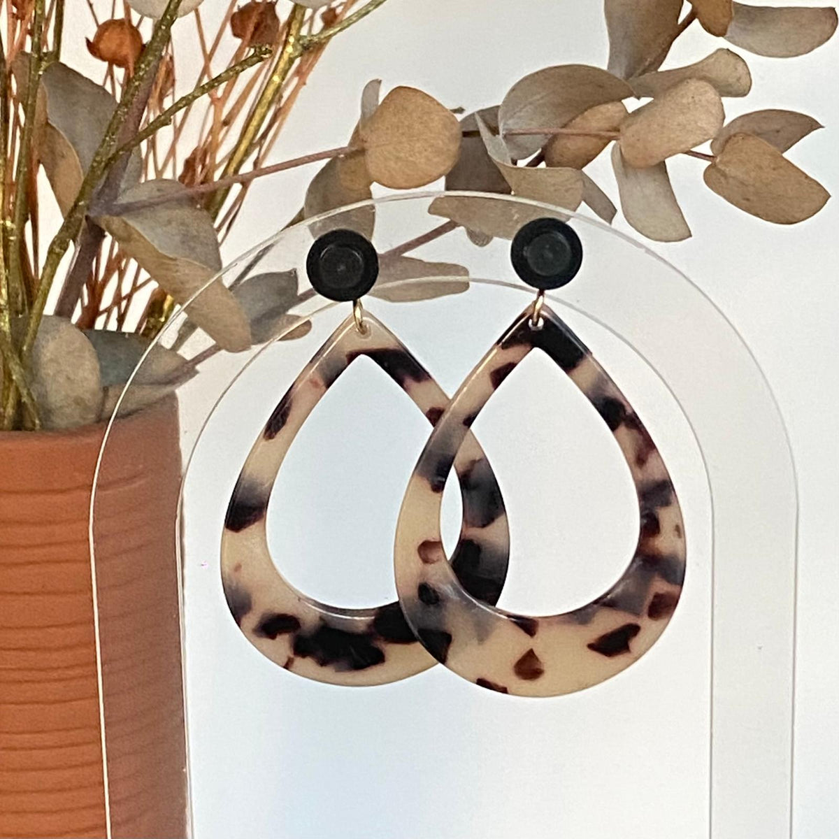 Boucles d’oreilles goutte en acétate écaille de tortue avec clou d’oreille en brique Lego noire, présentées sur un support transparent devant un bouquet sec.