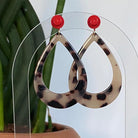 Boucles d’oreilles raffinée et tendance en forme de goutte en acétate au motif  écaille de tortue avec clou d’oreille en brique Lego rouge, présentées sur un support transparent devant un cactus.