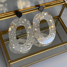 Boucles d'oreille paillettes or et Lego avec pièce ovale en acétate et éclats dorés, présentation sur une boîte à bijoux en verre.