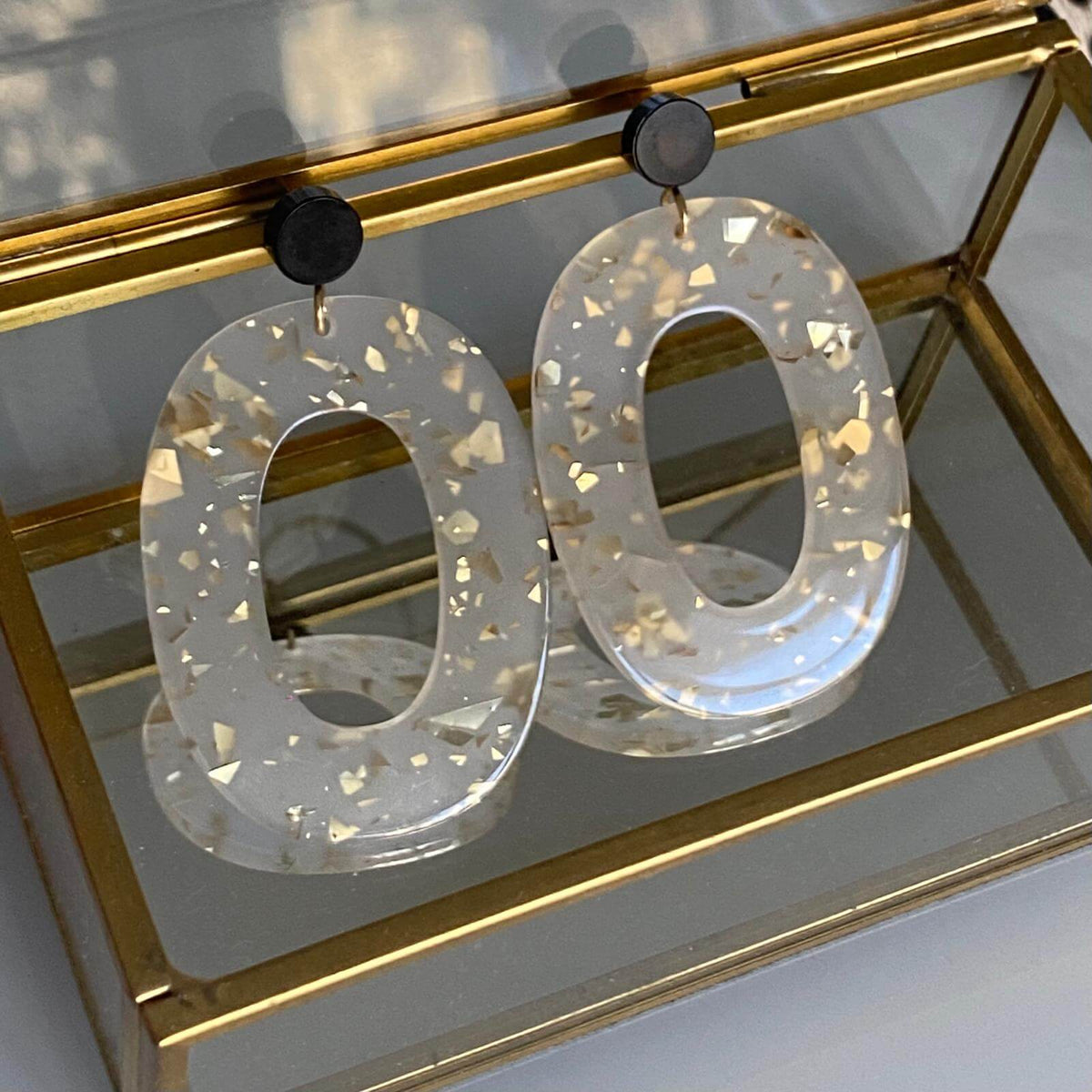 Boucles d'oreille paillettes or et Lego avec pièce ovale en acétate et éclats dorés, présentation sur une boîte à bijoux en verre.