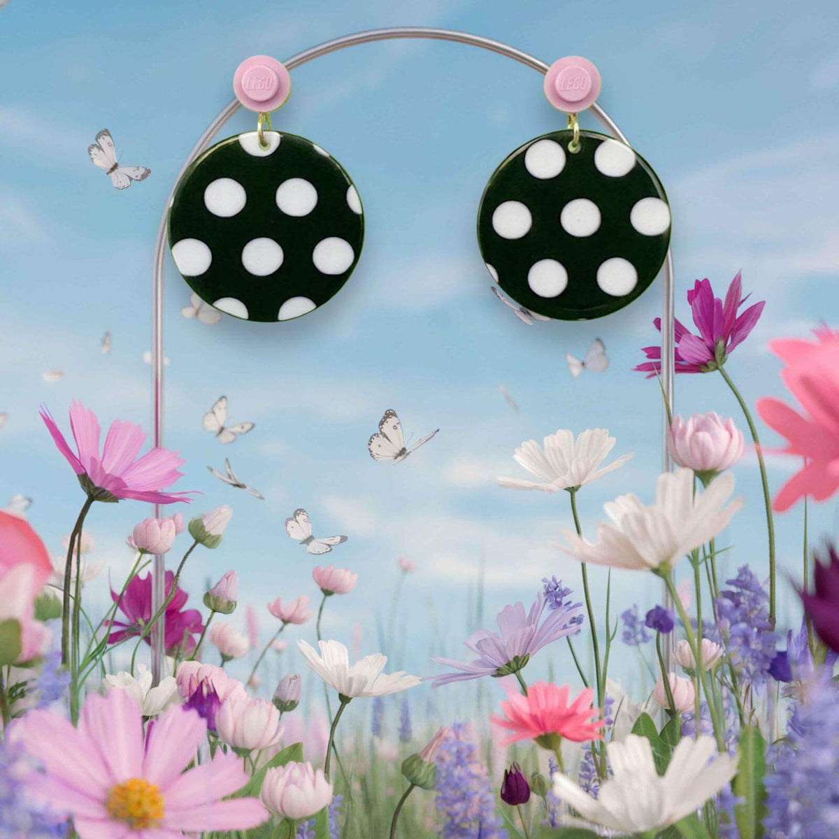 Boucles d'oreilles à pois noirs et blancs accrochées à l'oreille par une brique de Lego ronde rose pastel au milieu d'un décor féérique floral