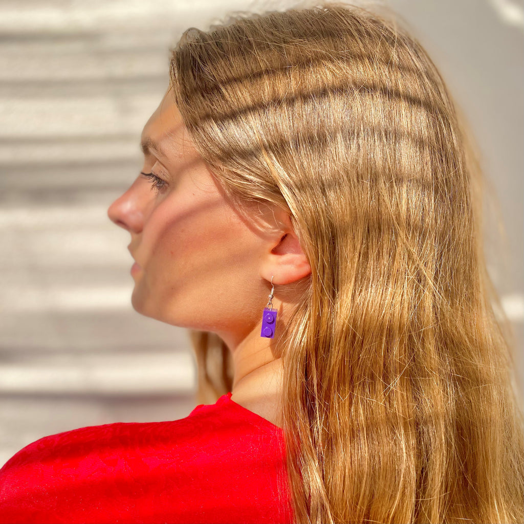 Femme portant des boucles d'oreilles pendantes en brique Lego violette, cheveux longs et blonds, vêtue d'un haut rouge.