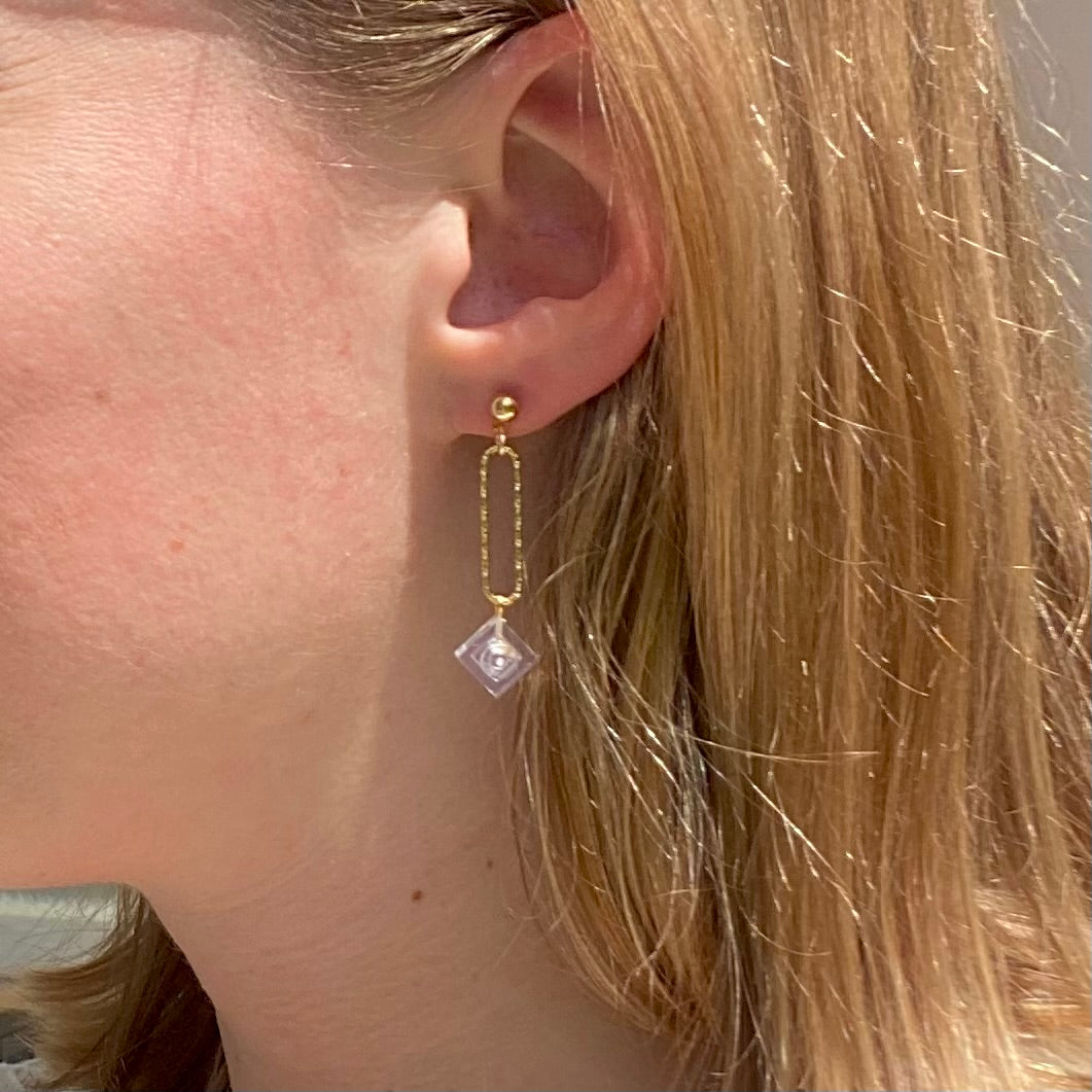 Boucle d'oreilles pendante accrochée à l'oreille par un clou d'oreille en forme de petite boule auquel est suspendu un anneau doré ovale martelé et en-dessous une brique de Lego 1x1 transparente comme du cristal. La boucle d'oreille est portée par une femme blanche blonde.