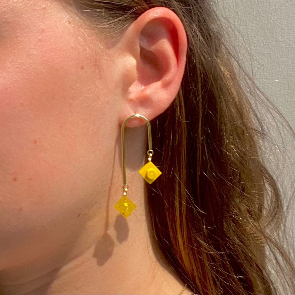 Boucles d'oreilles pendantes inspirées de l'art déco, avec briques Lego jaune suspendues, présentant un design graphique et asymétrique.