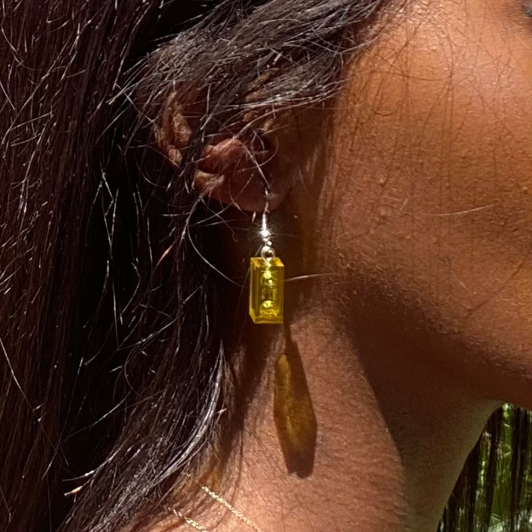 Boucles d'oreilles pendantes en brique Lego® jaune, ajoutant une touche colorée au look, portées avec des cheveux longs.
