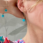 Boucle d'oreilles pendante accrochée à l'oreille par un clou d'oreille en forme de petite boule auquel est suspendu un anneau doré ovale martelé et en-dessous une brique de Lego 1x1 bleu turquoise. La boucle d'oreille est portée par une femme blanche blonde. Elle porte le bracelet Brique d'Oreille "Rockeuses" assorti.