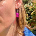 Boucles d'oreilles pendantes argentées avec brique Lego® rose accrochée à l'oreille en arrière-plan fleuri.