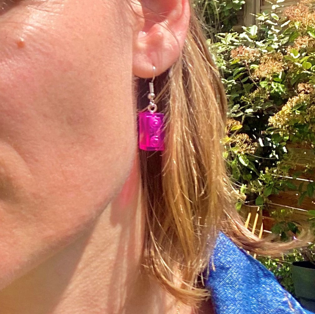 Boucles d'oreilles pendantes argentées avec brique Lego® rose accrochée à l'oreille en arrière-plan fleuri.