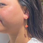 Boucle d'oreilles pendante accrochée à l'oreille par un clou d'oreille en forme de petite boule auquel est suspendu un anneau doré ovale martelé et en-dessous une brique de Lego 1x1 rose corail. La boucle d'oreille est portée par une femme malgache brune 