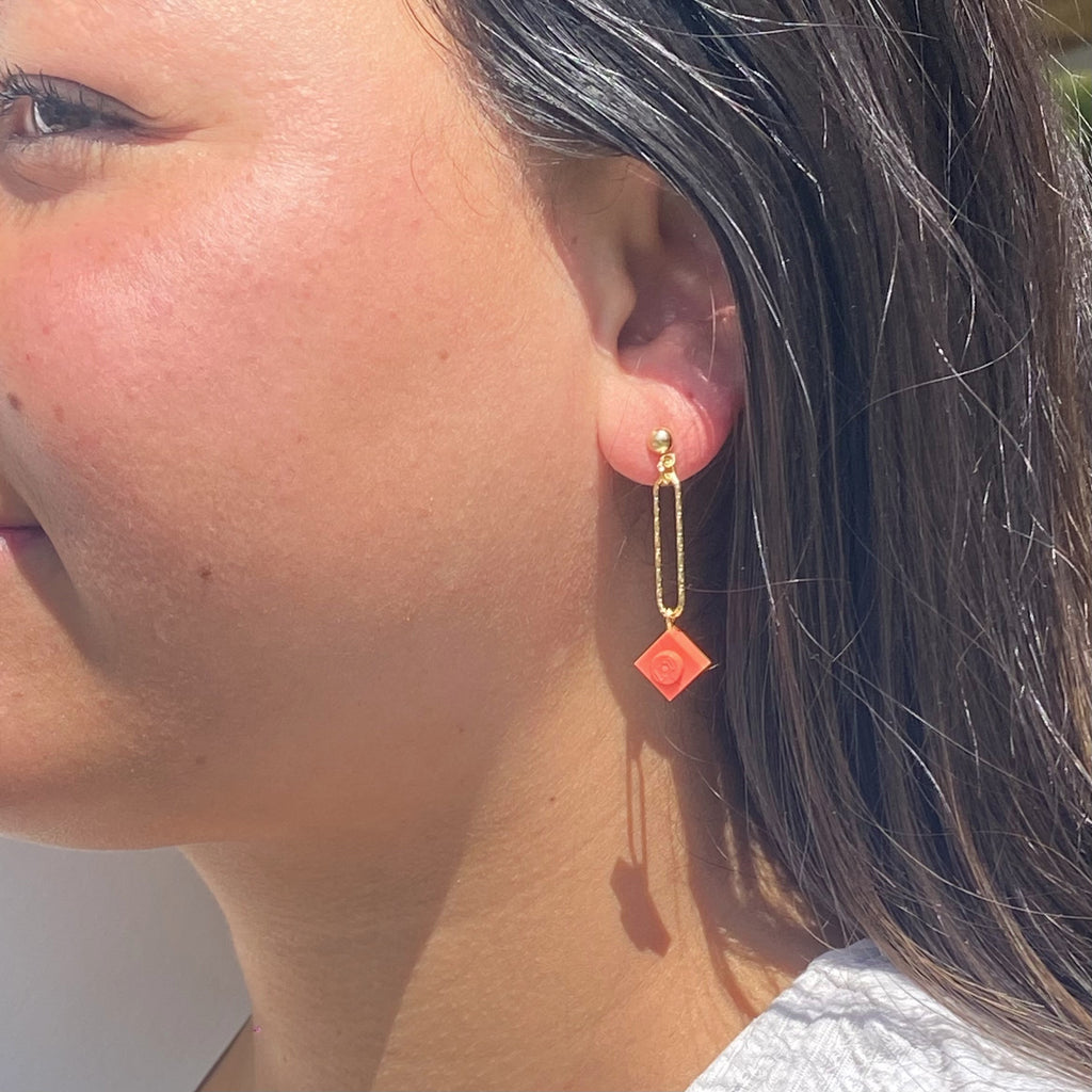 Boucle d'oreilles pendante accrochée à l'oreille par un clou d'oreille en forme de petite boule auquel est suspendu un anneau doré ovale martelé et en-dessous une brique de Lego 1x1 rose corail. La boucle d'oreille est portée par une femme malgache brune 