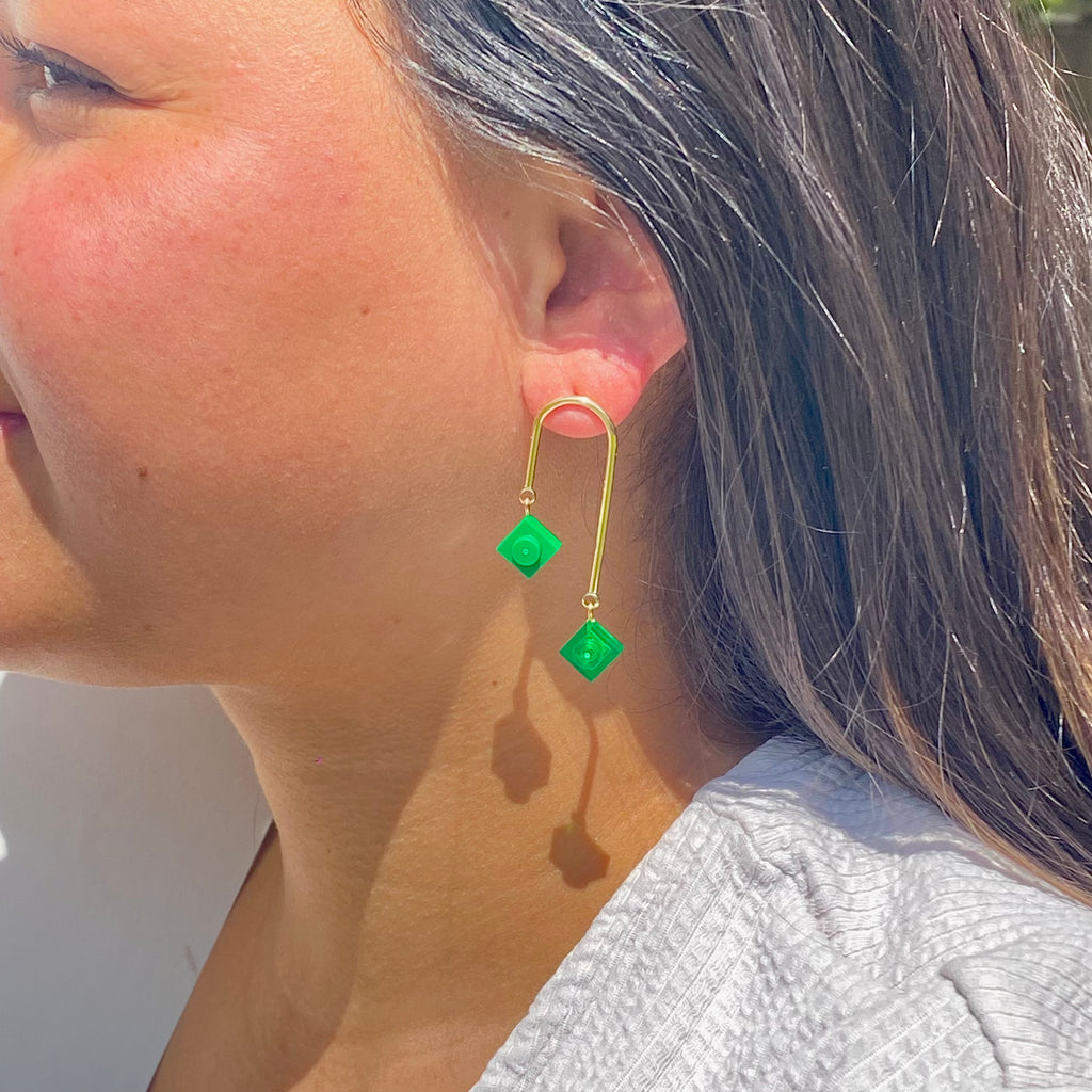 Boucles d'oreilles pendantes asymétriques avec 2 mini briques de LEGO vertes en style art déco, montrées sur un modèle féminin.