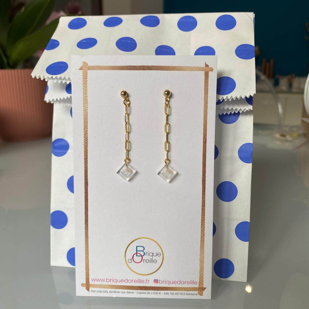Boucles d'oreilles pendantes dorées en chaîne avec briques Lego® transparentes, légères et colorées, présentées sur un emballage à pois.