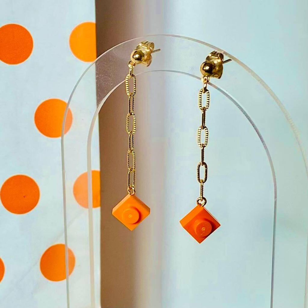 Boucles d'oreilles pendantes dorées avec briques Lego® orange sur un fond à pois colorés.