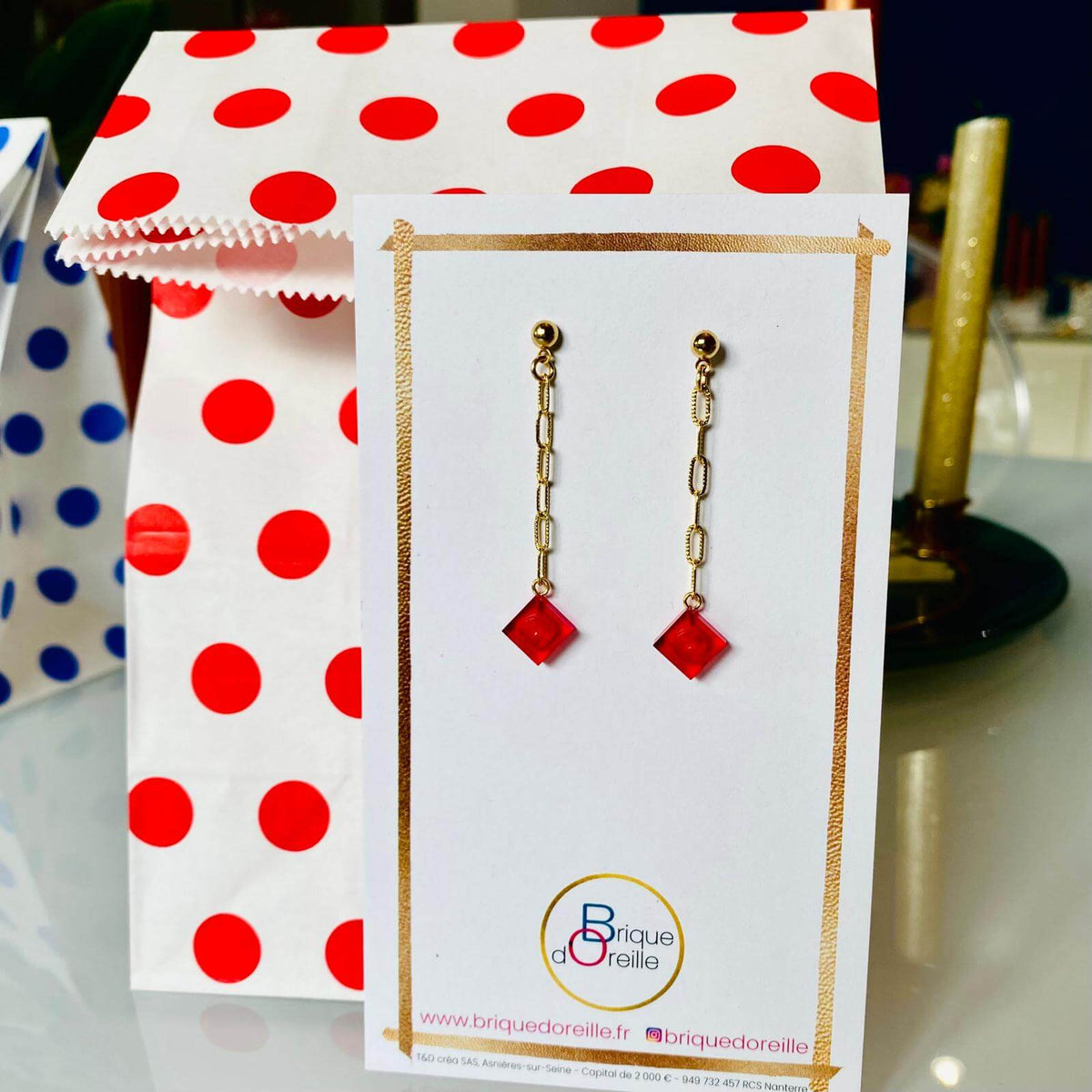 Boucles d'oreilles pendantes dorées avec briques Lego® rouge translucide effet rubis, présentées sur un support élégant.