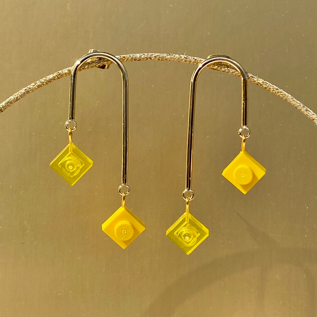 Boucles d'oreilles pendantes dorées avec petites briques Lego carrées jaunes, design asymétrique et graphique, inspirées de l'art déco.